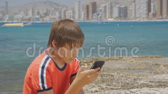 一幅英俊的少年用智能手机看着坐在摩天大楼旁边的码头上的屏幕的画像视频的预览图