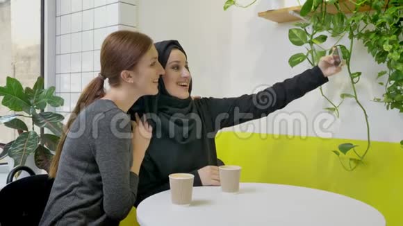 两个年轻漂亮的女人坐在咖啡馆里其中一个是戴着头巾的穆斯林用手机自拍看着电话视频的预览图