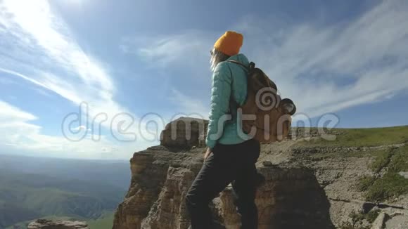 一个微笑的旅行女孩她背着背包戴着太阳镜拿着相机站在岩石高原的边缘视频的预览图