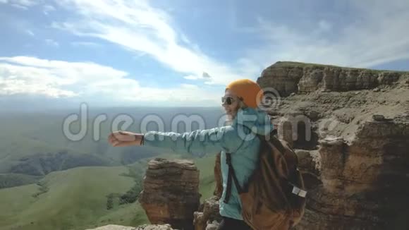 一个微笑的旅行女孩她背着背包戴着太阳镜拿着相机站在岩石高原的边缘视频的预览图