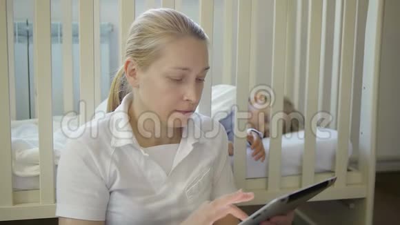 一个手里拿着电子平板电脑的女人坐在婴儿床上熟睡的小婴儿旁边视频的预览图