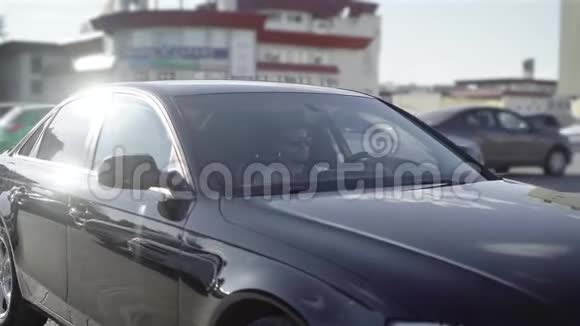 一个留着胡子的年轻人坐在他的黑色汽车上的停车场里商用轿车静静地沿着其他机器行驶视频的预览图