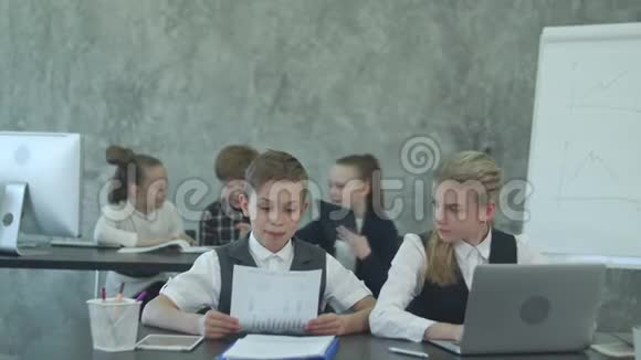 一个金发女孩和一个穿着西装的男孩坐在办公室的桌子上用笔记本电脑工作然后关闭它并写下它视频的预览图