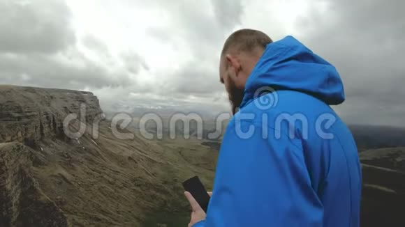 长胡子的嬉皮士男子在高加索山脉的岩石上用智能手机拍照视频的预览图