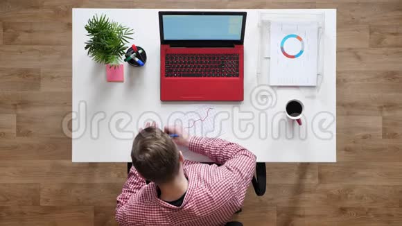 戴眼镜的年轻人工作写在纸上拍在上面坐在桌子后面拿着咖啡和笔记本电脑视频的预览图