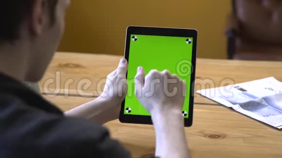 一位身穿黑色衬衫的年轻人坐在木桌旁端着平板电脑在彩色钥匙绿色屏幕上滑动视频的预览图