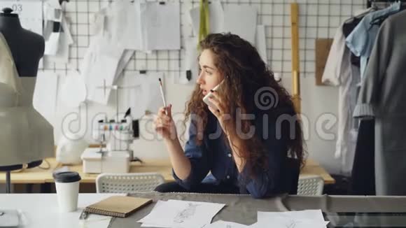 专业的时装设计师用手机说话用铅笔在桌子上画服装素描做裁缝视频的预览图