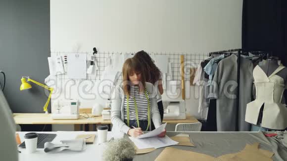 年轻女性的服装设计师正在画素描当她的同事带着草稿来找她时展示并改进了她的服装视频的预览图