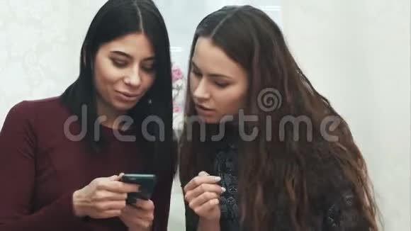 两个女孩用智能手机说话微笑坐在沙发上视频的预览图