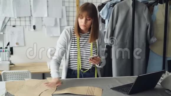 年轻女裁缝正在检查服装纸样并在看智能手机时用卷尺测量视频的预览图