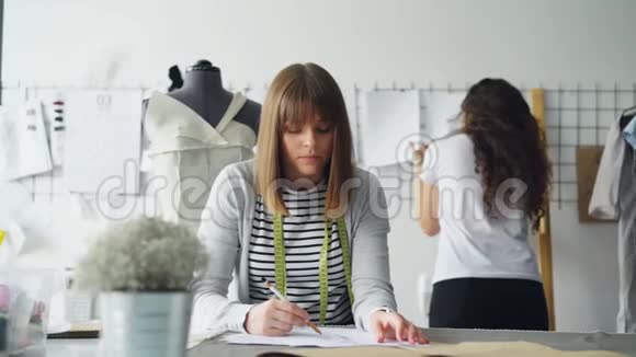 年轻的服装设计师忙着用铅笔画素描而她的同事正在看墙上钉着的画视频的预览图