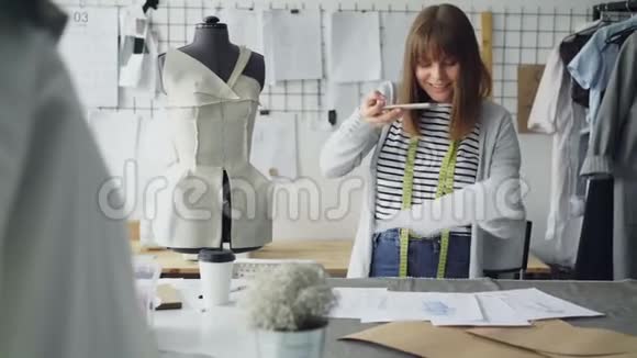 雄心勃勃的创意女裁缝将服装草图放在工作室办公桌上并用智能手机拍摄视频的预览图