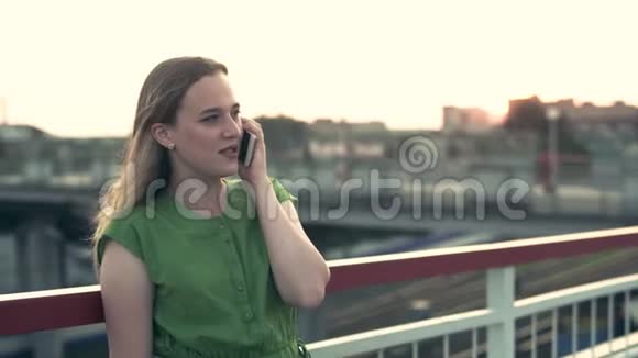 日落时分一个年轻的女孩坐在智能手机上穿着绿色的裙子笑着视频的预览图
