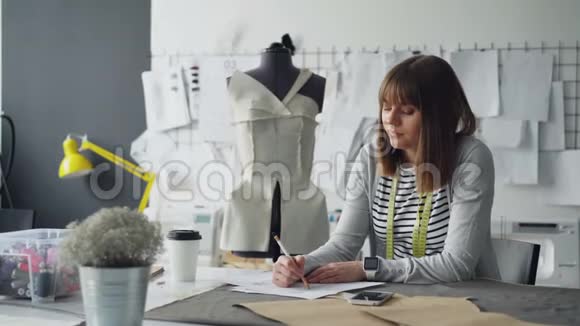 专业服装设计师正在绘制草图然后在智能手机上接收信息她在拿装置触摸屏幕视频的预览图