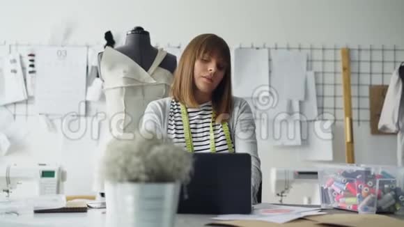 裁剪工作室的老板正在工作场所使用笔记本电脑而她的设计师正带着草图来找她妇女视频的预览图