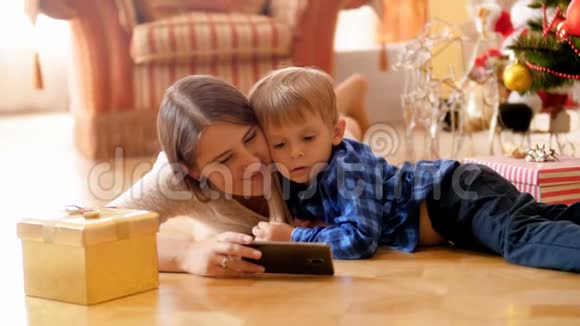 快乐的年轻妈妈在圣诞节和她的小儿子躺在花架上用智能手机视频的预览图
