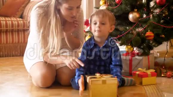 4k视频可爱的微笑幼儿男孩坐在圣诞树下与母亲和摇动礼品盒礼物视频的预览图