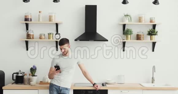 智能手机男人在家厨房的手机上浏览英俊的年轻人在智能手机上浏览微笑着快乐视频的预览图