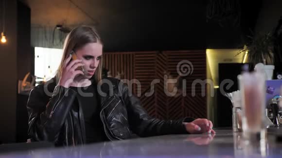 年轻漂亮的女人穿着黑色夹克浓妆艳抹坐着打电话愤怒酒吧背景视频的预览图