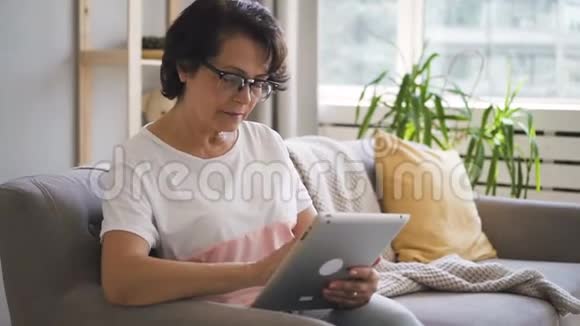 成熟的家庭主妇使用平板电脑坐在沙发上的现代家庭内部视频的预览图
