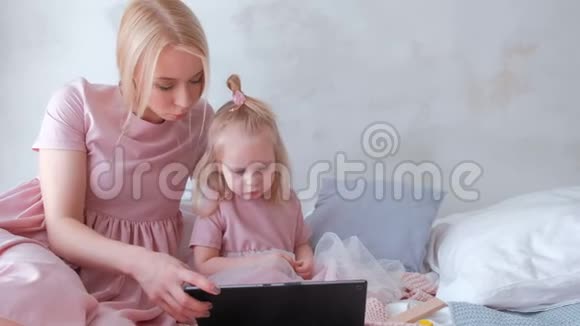 年轻漂亮的金发女郎穿着粉色连衣裙的迷人女儿在平板电脑上观看视频视频的预览图