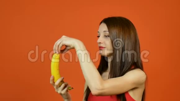 年轻迷人的女人手里拿着黄色香蕉然后在明亮的橙色背景下吃它热带水果视频的预览图