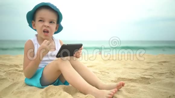 可爱的男孩坐在沙滩上使用智能手机应用程序视频的预览图