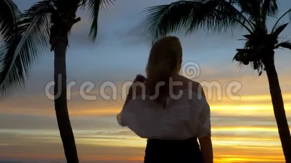 日落时分金发女孩站在海滩上的棕榈树旁视频的预览图