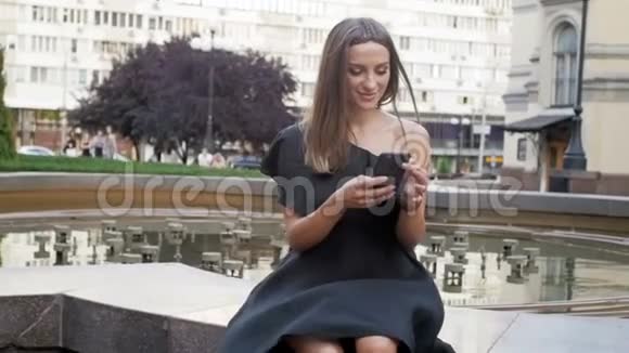 在大风天穿着短裙的年轻女子坐在公园里用智能手机打字的慢镜头视频的预览图