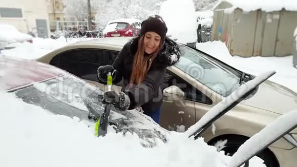 4k视频美丽微笑的年轻女子用伸缩刷子从雪中清洁汽车视频的预览图