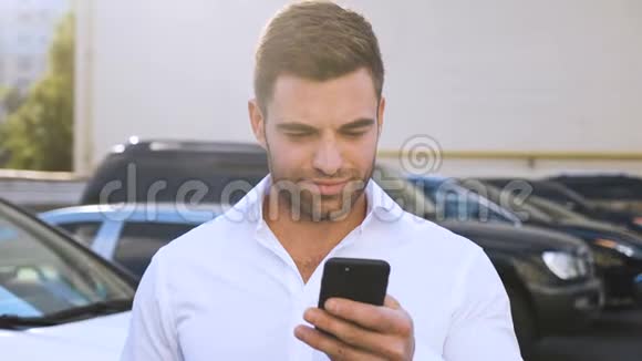 站在办公楼附近的停车场上用智能手机打字的年轻帅哥英俊的商人视频的预览图
