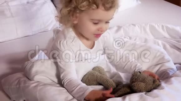 可爱的小女孩亲吻她最好的朋友喜欢的玩具泰迪熊坐在白色的床上视频的预览图