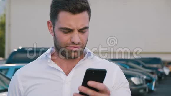 站在办公楼附近的停车场上用智能手机打字的年轻帅哥英俊的商人视频的预览图