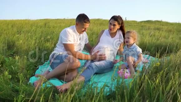 一对怀孕的夫妇和一个蹒跚学步的女儿在草地上的毯子上休闲视频的预览图