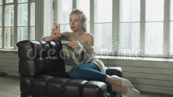 年轻的美女正在用信用卡通过电话购买金发女郎坐在家里的扶手椅上视频的预览图