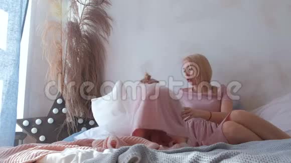 年轻漂亮的金发妈妈和穿着粉红色连衣裙的可爱女儿躺在床上玩耍视频的预览图