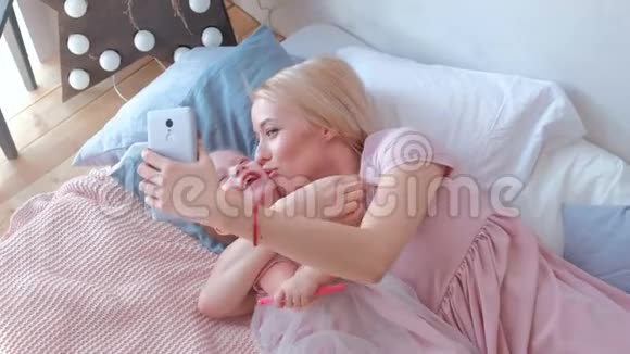 年轻迷人的金发妈妈和她穿着粉红色连衣裙的小迷人女儿在手机上拍照家庭拥抱视频的预览图