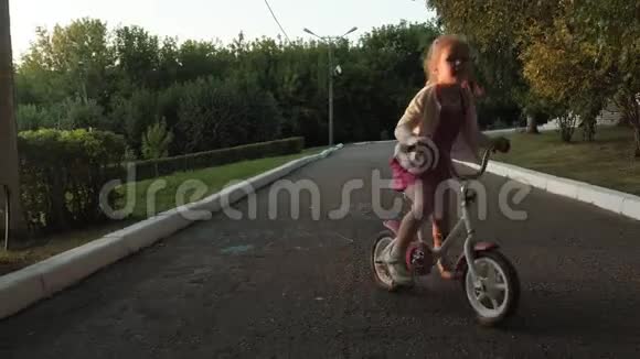 一个快乐美丽金发碧眼的小女孩穿着粉红色的裙子跳投骑着一辆儿童自行车上路视频的预览图