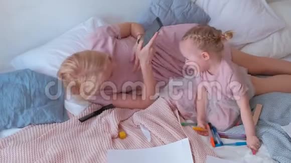 年轻迷人的金发妈妈躺在床上她的小可爱女儿穿着粉红色的连衣裙和手机视频的预览图
