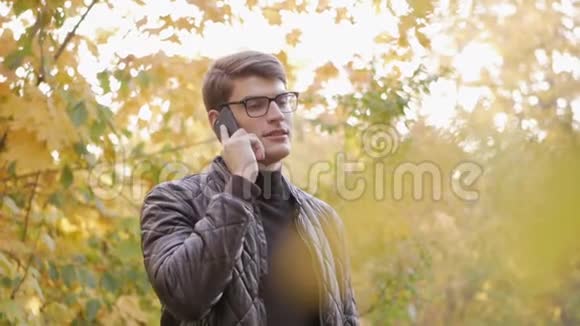阳光明媚的一天一个年轻有魅力的男人打着电话在秋天公园散步视频的预览图