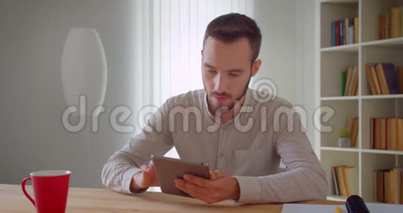 年轻英俊的白种人商人在公寓室内平板电脑上浏览的特写照片视频的预览图
