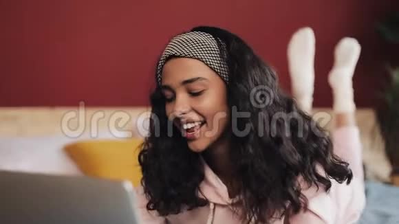 快乐的非裔美国女人躺在床上用笔记本电脑浏览网页穿着粉色睡衣的女孩微笑着视频的预览图