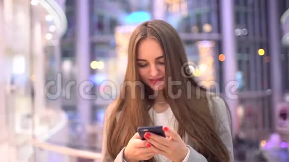 美丽的快乐女孩长发在智能手机上写着一条信息站在商场里4k视频的预览图