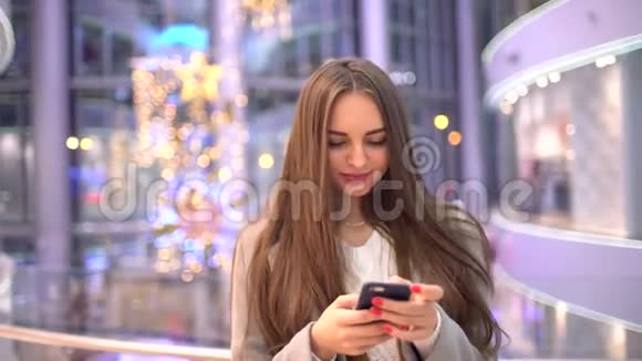 年轻女子微笑着浏览互联网或短信短信使用现代智能手机在模糊的大商场背景4视频的预览图
