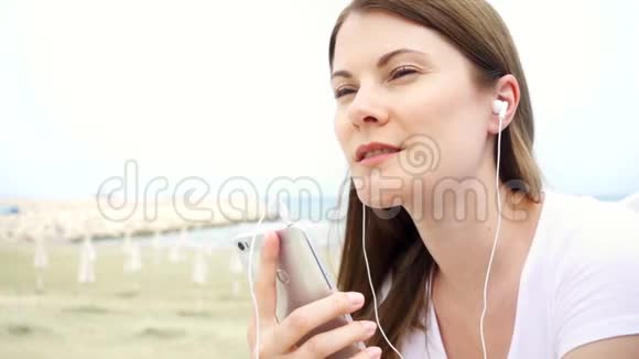 年轻微笑的女人独自在海滩上使用手机女性通过手机慢动作听音乐视频的预览图