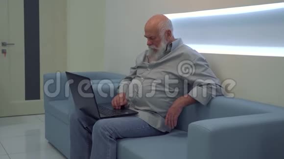 一个成熟的男人坐在沙发上一边用笔记本电脑一边在诊所等候视频的预览图