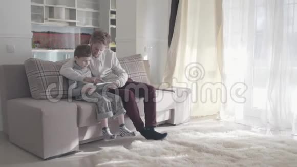 爸爸和儿子用数码平板电脑坐在大客厅的沙发上幸福的家庭一起度过时光爸爸视频的预览图