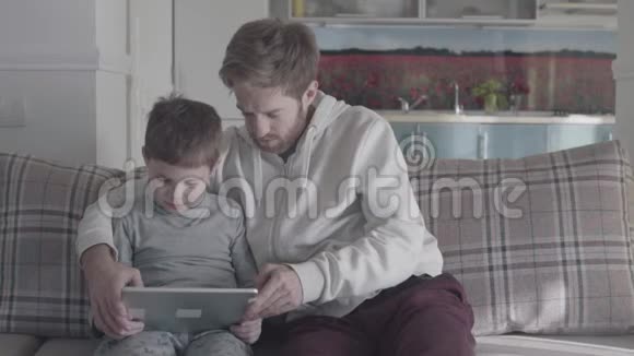 父子俩坐在大客厅的沙发上玩平板电脑爸爸教他的孩子快乐的家庭开支视频的预览图