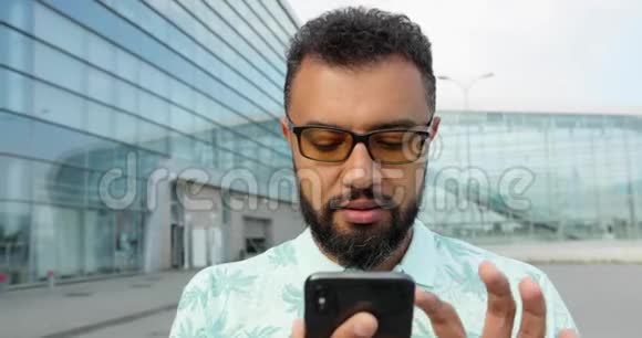 这位戴眼镜的自信严肃的非洲男子用手机在玻璃建筑上的特写肖像他是视频的预览图