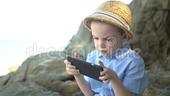 可爱的男孩坐在石头上使用智能手机在海边看手机应用程序视频的预览图